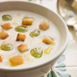 Ajo blanco adornado con uvas y picatostes presentado en cuenco de cerámica junto a cuchara