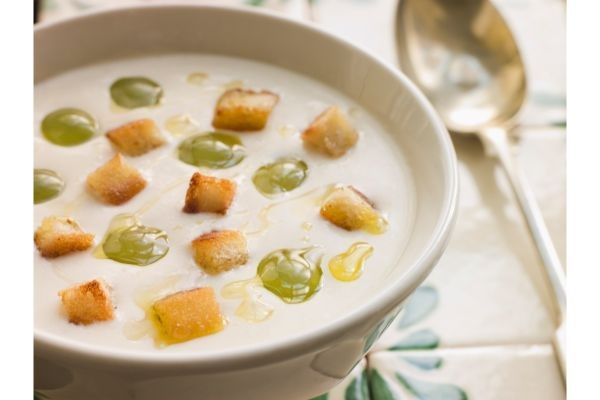 Ajo blanco adornado con uvas y picatostes presentado en cuenco de cerámica junto a cuchara