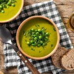 Crema de guisantes presentada en cuenco decorada con menta y guisantes con cuchara y pan alrededor