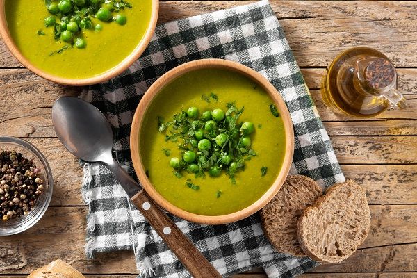 Crema de guisantes presentada en cuenco decorada con menta y guisantes con cuchara y pan alrededor