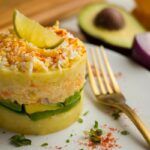 Causa limeña presentada en plato blanco con tenedor e ingredientes detrás
