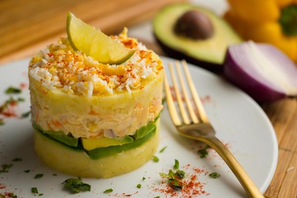 Causa limeña presentada en plato blanco con tenedor e ingredientes detrás