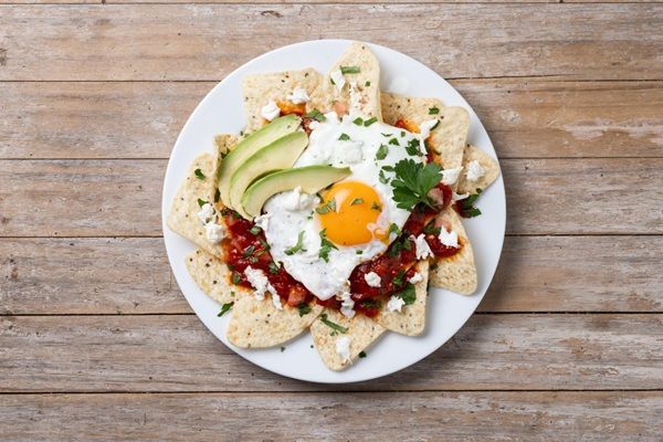 Chilaquiles rojos mexicanos con huevo, pollo y aguacate presentados en plato blanco sobre mesa de madera vista desde arriba