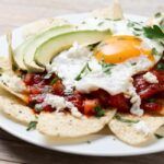 Chilaquiles rojos mexicanos con huevo, pollo y aguacate presentados en plato blanco sobre mesa de madera vista de lado