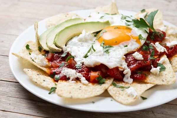 Chilaquiles rojos mexicanos con huevo, pollo y aguacate presentados en plato blanco sobre mesa de madera vista de lado