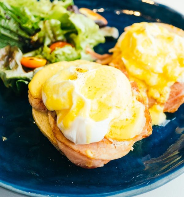 Huevos benedict con muffin inglés, jamón y salsa holandesa presentados en plato junto a ensalada vista de cerca