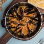 Mejillones a la marinera receta catalana presentados en sartén sobre tabla de madera junto a pan tostado
