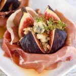 Primer plano de higo relleno con queso azul envueltos en jamón y con miel presentado en planto blanco cuadrado