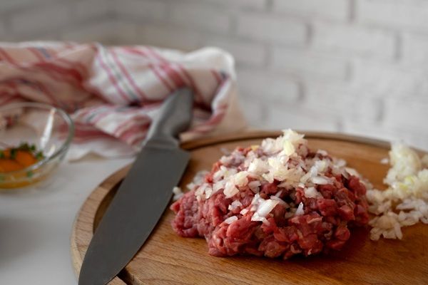 Carne mezclada con cebolla y otros ingredientes encima de tabla de madera junto a cuchillo