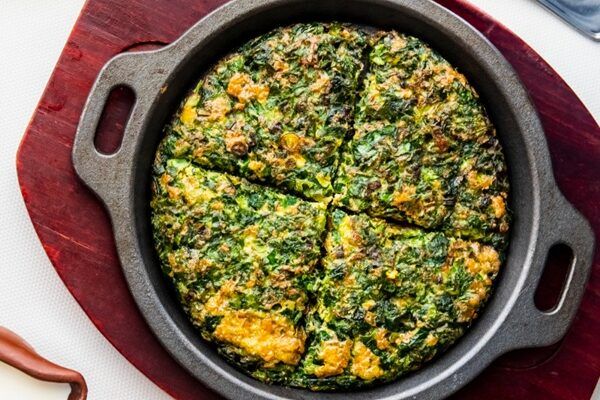 Tortilla persa de hierbas o kuku sabzi cortada en cuatro trozos presentada en sartén negra sobre tabla de madera
