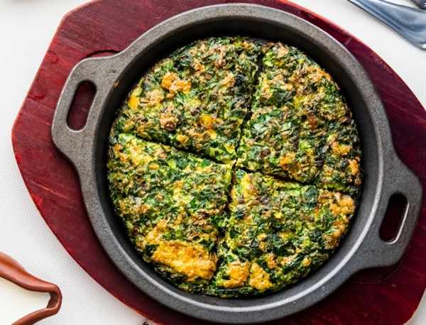 Tortilla persa de hierbas o kuku sabzi cortada en cuatro trozos presentada en sartén negra sobre tabla de madera