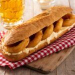Bocadillo de calamares con mayonesa sobre paño a cuadros rojo, tabla de madera y cerveza al fondo